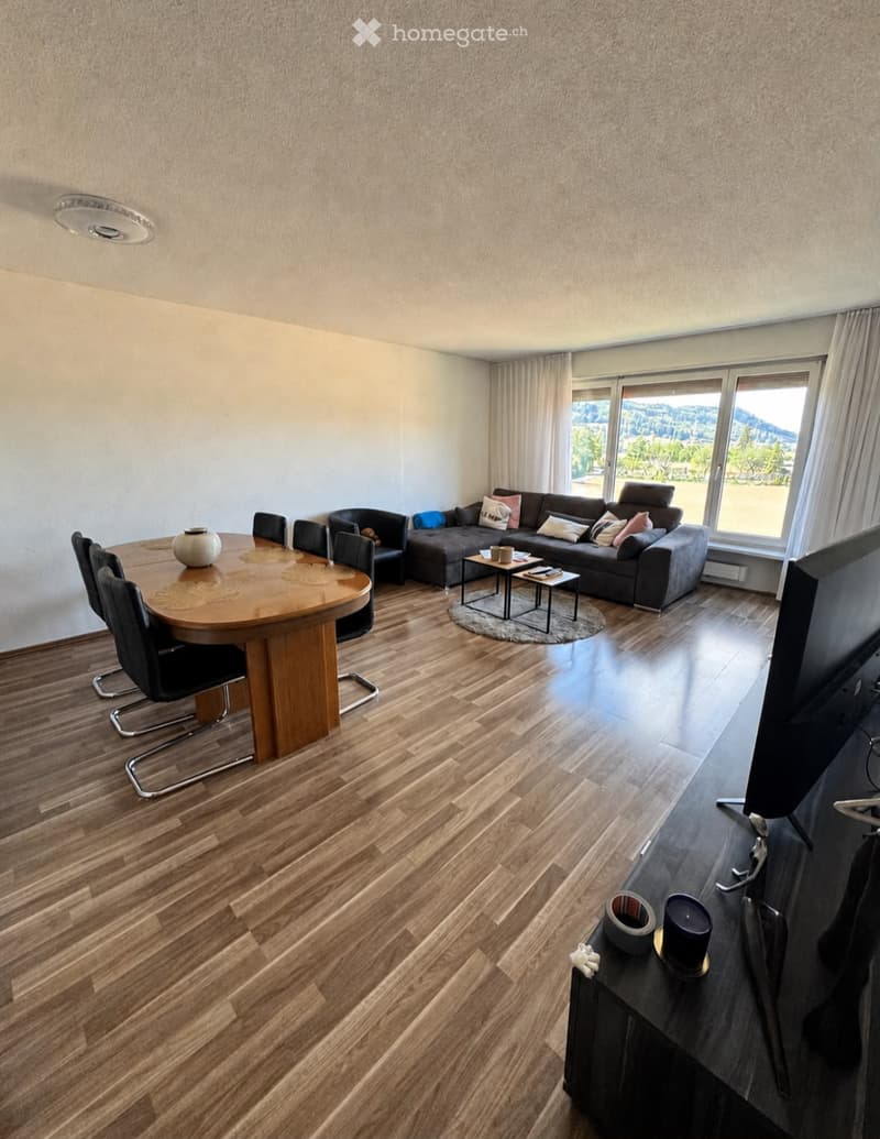 Spacious living room with wooden flooring, large windows, grey sofa, coffee table, dining table, chairs, and TV stand.