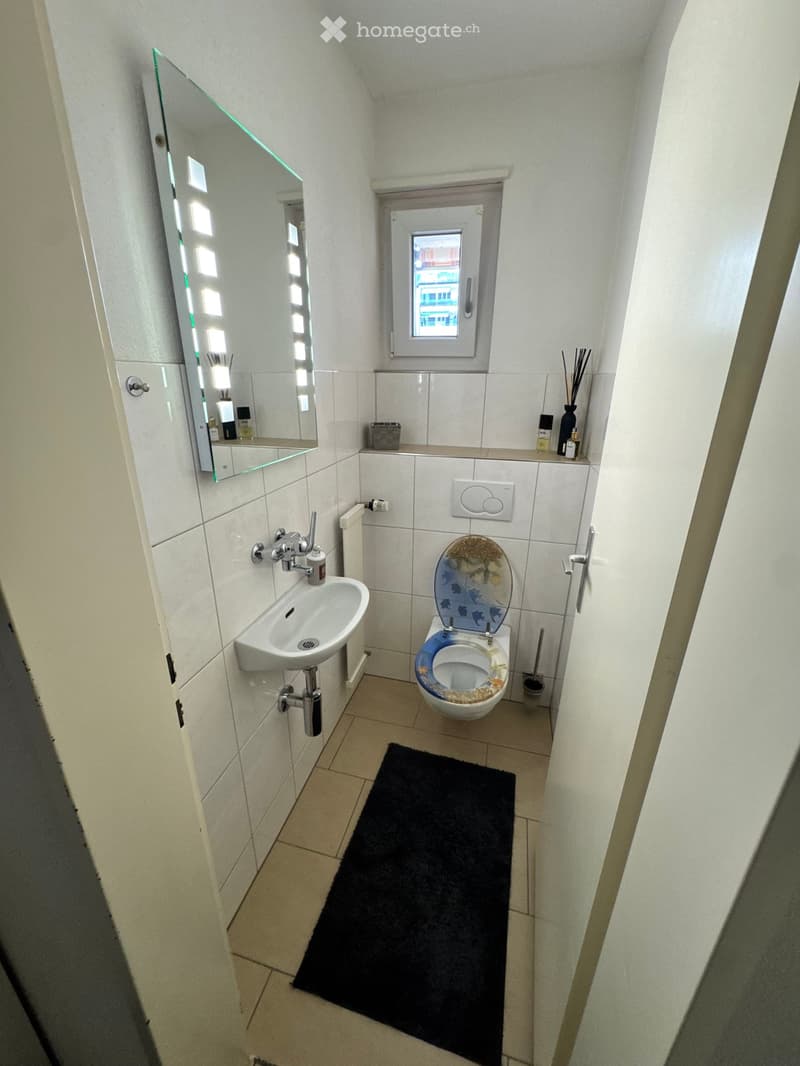Modern bathroom with white tiled walls, a toilet with a decorative seat cover, a sink with a modern faucet, a large mirror with built-in lighting, a black bathroom rug, and a small window providing natural light.