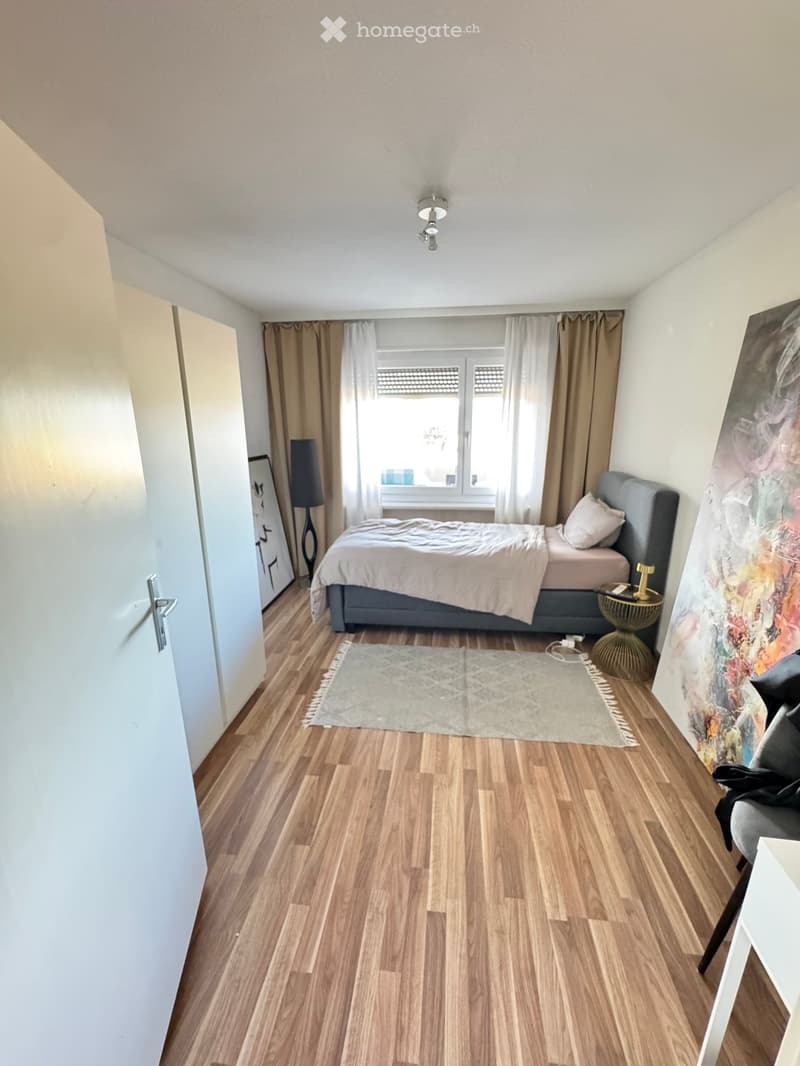 Cozy bedroom with wooden flooring, a grey upholstered bed, a nightstand with a lamp, a small round side table, a rug, and large windows with curtains.