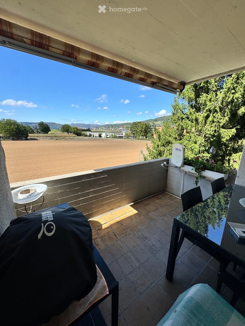 Balcony with tiled floor, outdoor furniture including a table, chairs, and a covered barbecue, overlooking a scenic countryside view.