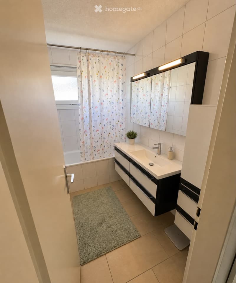 Modern bathroom with a white tiled wall, a black vanity with a sink, a mirror with built-in lighting, a bathtub with a floral shower curtain, a small potted plant, and a grey rug on the floor.