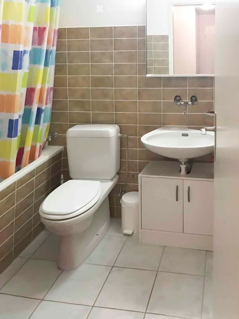 Bathroom with a toilet, sink, bathtub, and a small cabinet. The walls are tiled in brown and beige, and the floor is tiled in light gray.