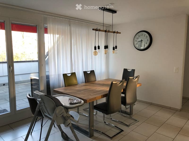 Dining room with a wooden dining table, six chairs, a baby chair, and a wall clock. Large windows with white curtains lead to a balcony.