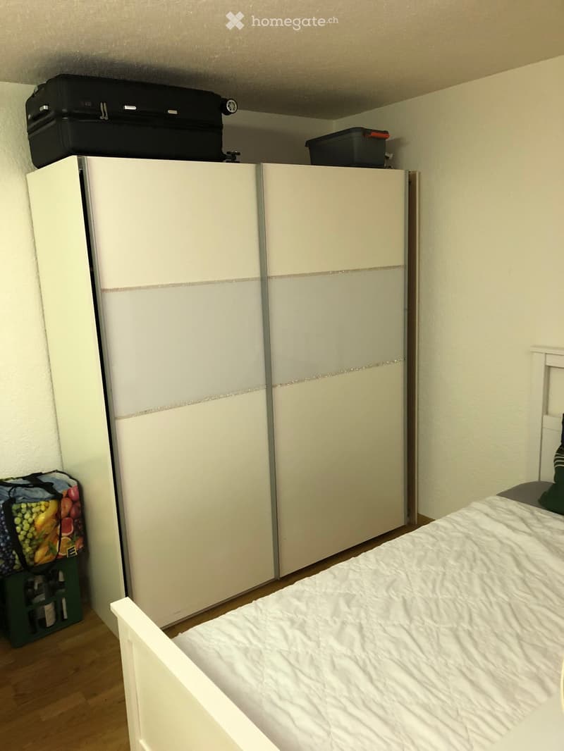 A tidy bedroom with a white bed, a large wardrobe, and a suitcase on top. A fruit basket is placed on the floor near the wardrobe.