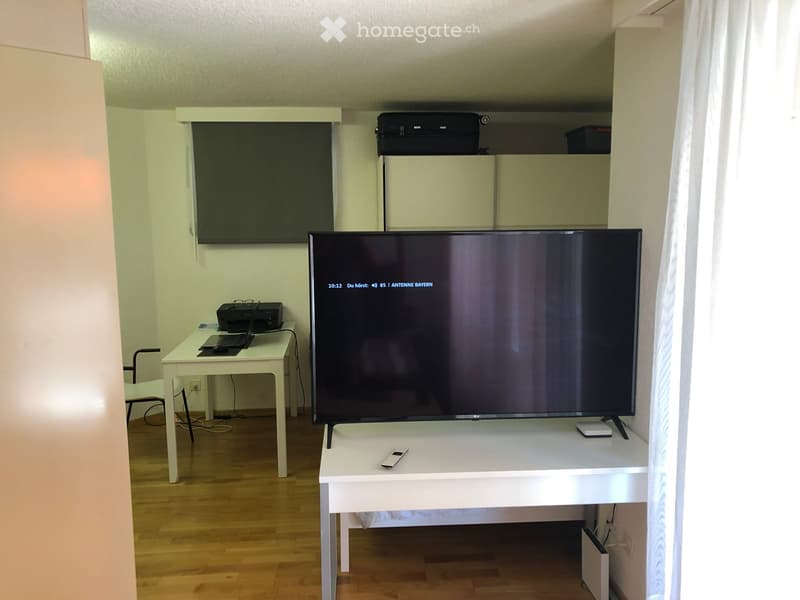 A modern living room with a large flat-screen TV on a white stand, a small white table with a laptop and printer, and a chair near the table.