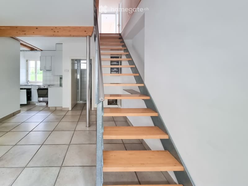 Modern hallway with a wooden staircase, metal railing, and tiled floor. The staircase leads to an upper level, with a kitchen visible in the background.