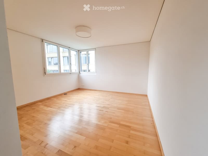 Empty room with light wooden flooring, white walls, and large windows allowing natural light. The room features a modern, minimalist design with a recessed ceiling light fixture.