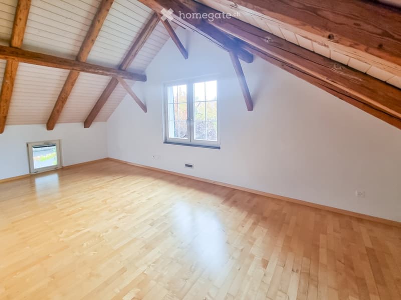 Empty attic room with wooden beams, light wooden flooring, and large windows allowing natural light to enter.
