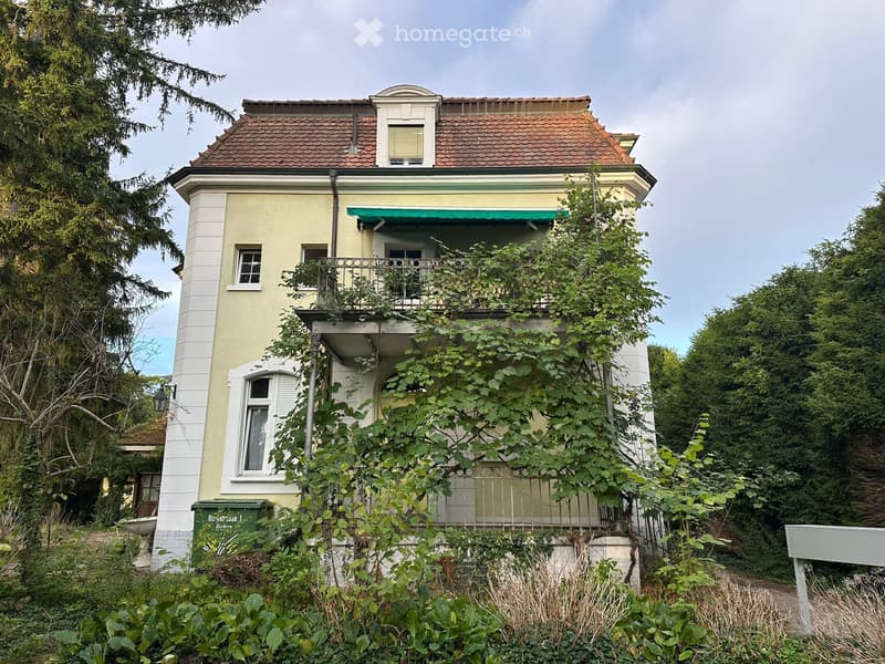 Two-story house with a balcony, surrounded by greenery and trees.