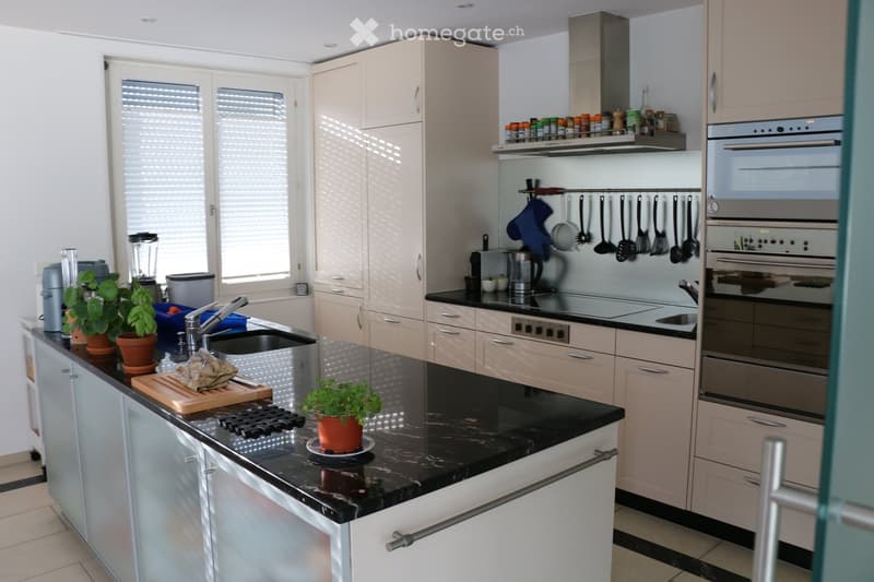 Modern kitchen with light-colored cabinets, black countertops, stainless steel appliances, and a central island with a sink and cooking area.