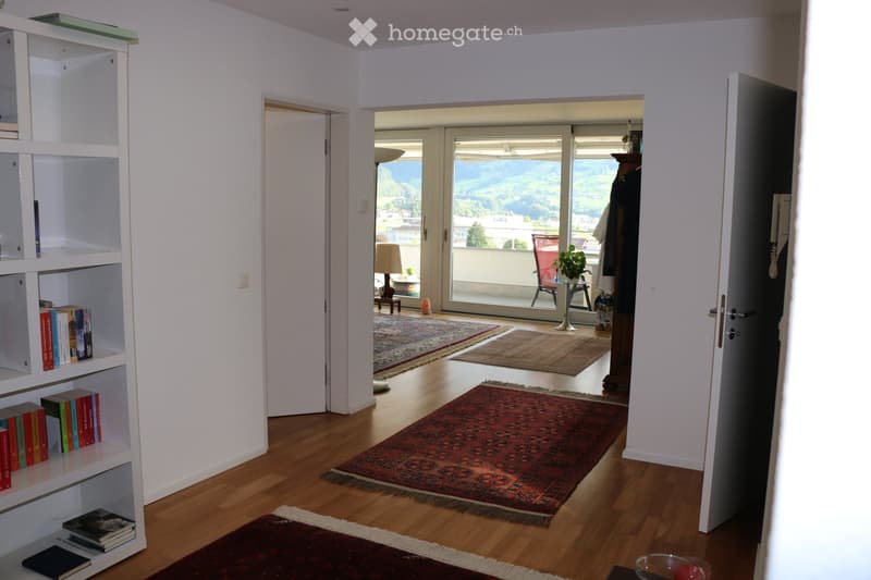 Bright hallway with wooden flooring, white walls, and a large bookshelf filled with books. A red rug lies on the floor, and a doorway leads to a balcony with a scenic mountain view. A small table and chair are visible on the balcony.