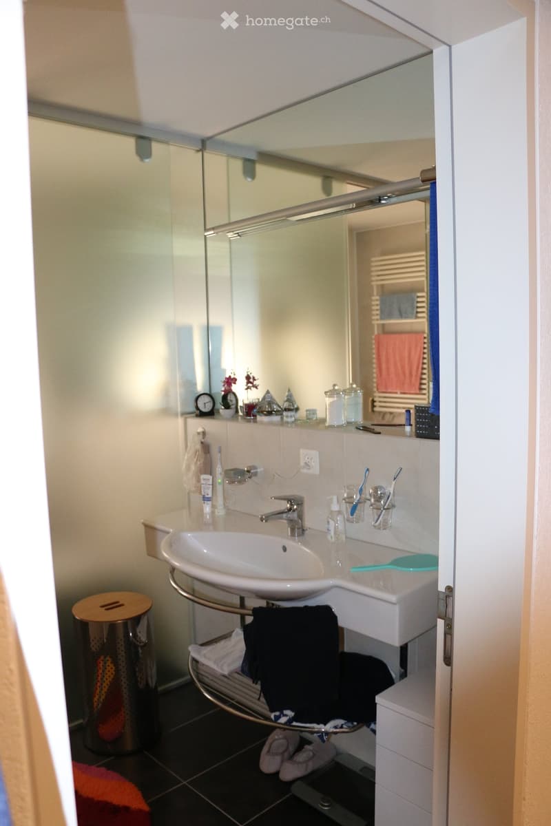 Modern bathroom with a white sink, chrome faucet, large mirror, and various toiletries on the counter. A towel rack holds a black towel and a white towel, and a trash can is visible in the corner.