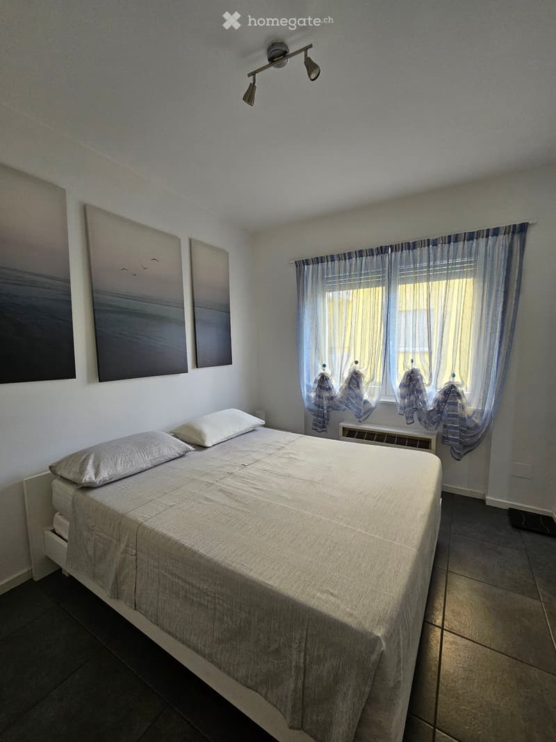 A furnished bedroom with a single bed, pillows, a radiator, and two paintings on the wall. The room has tiled flooring and a window with sheer curtains.