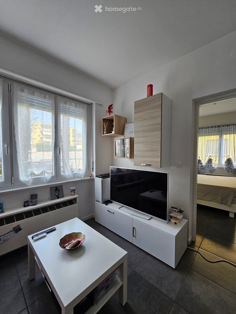 A modern living room with a white table, a large flat-screen TV on a white stand, and a small cabinet with a mirror. The room has large windows with white curtains and a view of the outside.