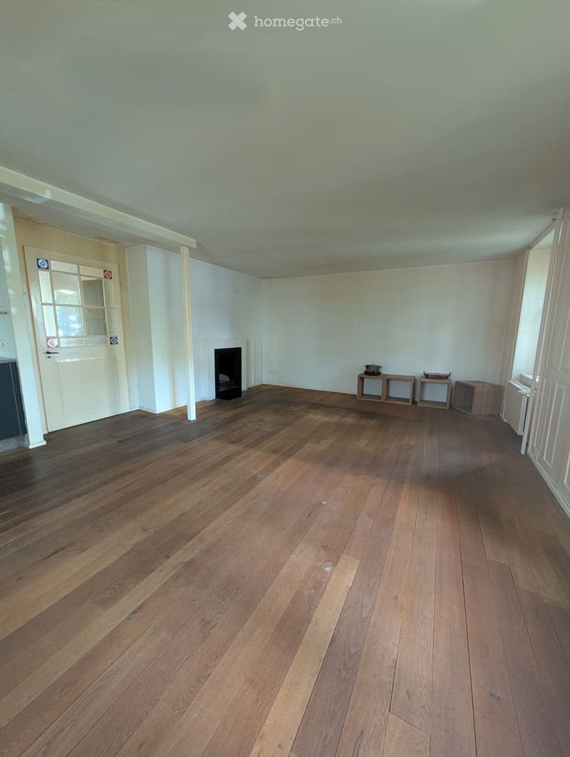 Empty room with wooden flooring, white walls, and a fireplace. There are small wooden tables and a radiator visible.