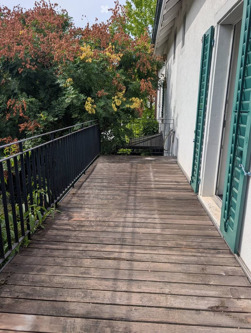 Wooden balcony with black metal railing, green shutters, and lush greenery in the background.
