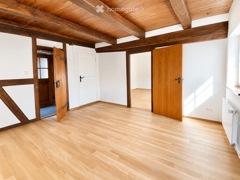 Empty room with wooden flooring, exposed wooden beams on the ceiling, and several doors leading to other spaces.