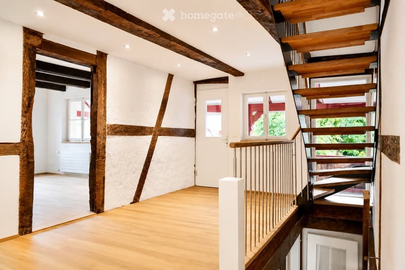Empty room with wooden floor, exposed wooden beams, and a staircase leading upwards. The space is well-lit with recessed lighting and has white walls with a textured finish. There are windows on one side and a door leading to another area.