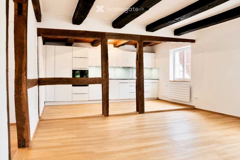 Empty room with wooden floor, white walls, exposed wooden beams, white kitchen cabinets, and a window with a view of a tiled roof.