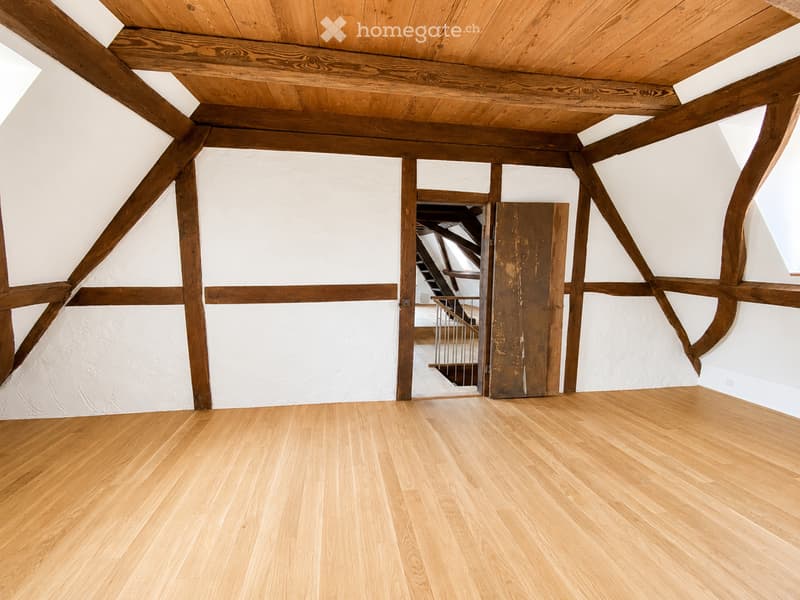 Empty room with wooden floor and exposed wooden beams on the ceiling and walls. A partially open wooden door leads to a staircase area.