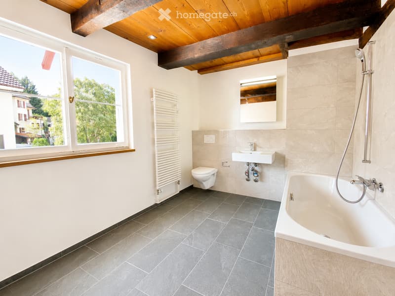 Modern bathroom with a bathtub, toilet, sink, radiator, and a mirror. The room features a wooden ceiling with exposed beams and large windows offering a view of the outdoors.