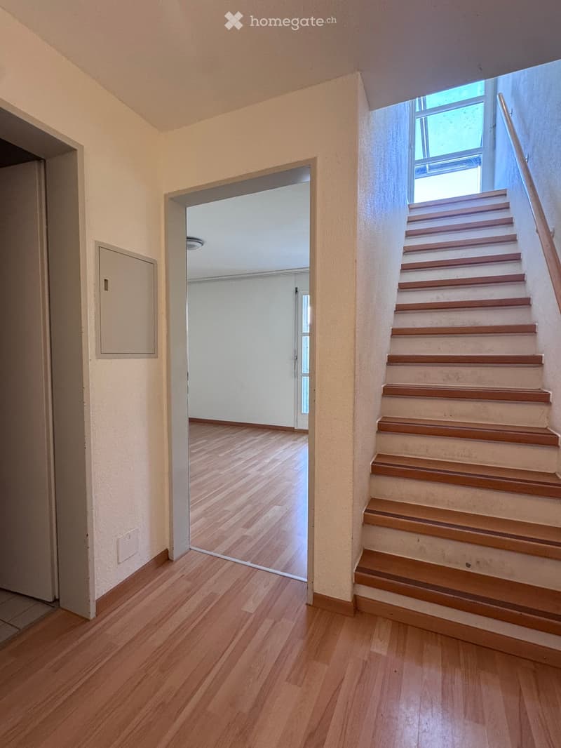 A clean, well-lit hallway with wooden flooring, a staircase leading upwards, and an open doorway to another room.