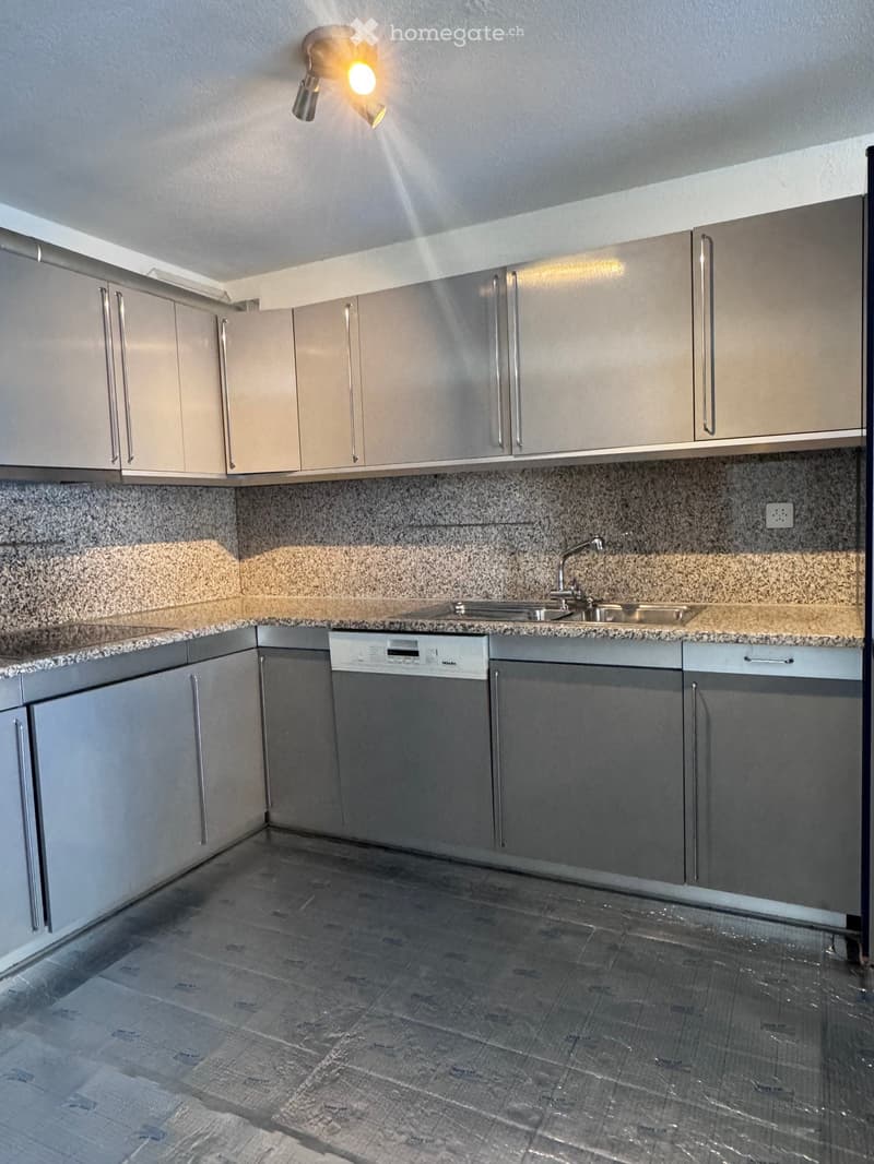 Modern kitchen with grey cabinets, granite countertops, and a built-in dishwasher. The floor is tiled in a dark, polished finish.