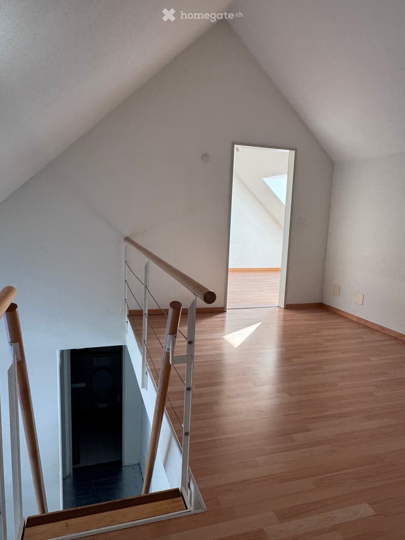 Spacious hallway with wooden flooring, modern staircase featuring a wooden handrail and metal balustrade, leading to an open doorway with natural light.
