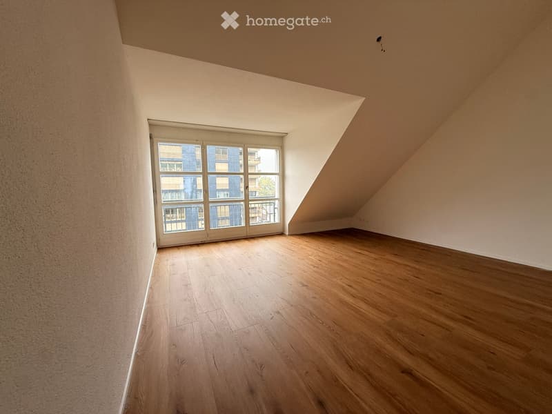 Empty room with wooden flooring and large windows, featuring a slanted ceiling and neutral-colored walls.