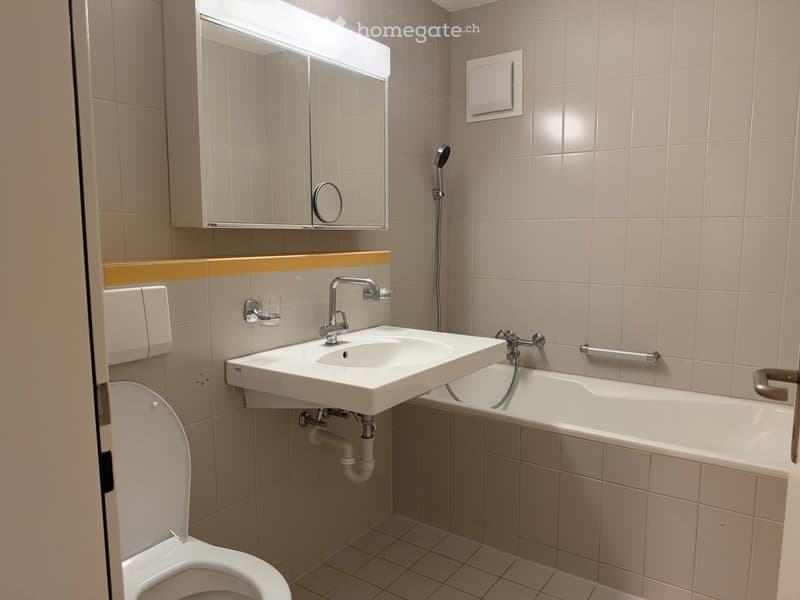 Modern bathroom with a white toilet, sink, bathtub, mirror, and showerhead. Light beige tiles and a yellow accent stripe above the sink.