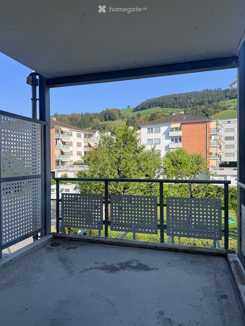 Balcony with metal railing and perforated panels, overlooking a residential area with trees and hills in the background.