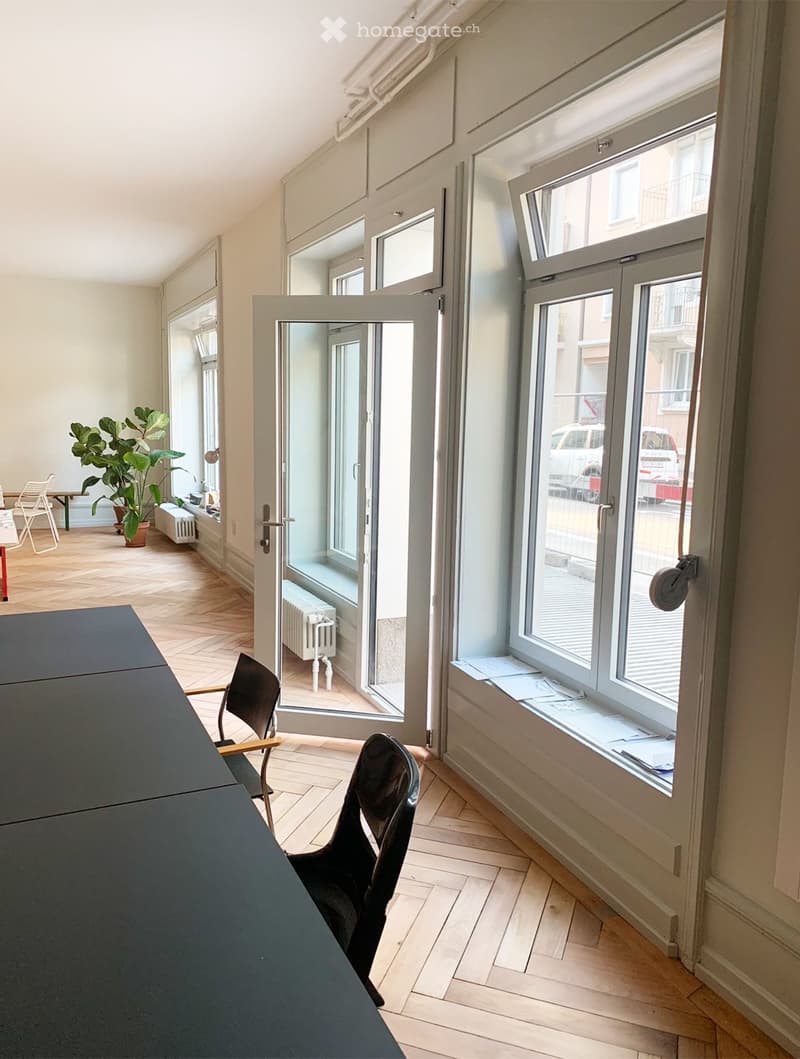 Bright, open room with wooden flooring, large windows, and a sliding glass door leading to a balcony. Includes a black table, two black chairs, a small table with chairs, and a potted plant in the corner.
