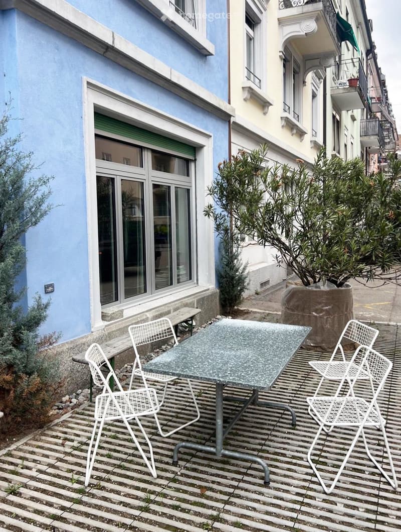 Balcony with a glass table and four white metal chairs, surrounded by plants and a stone wall.
