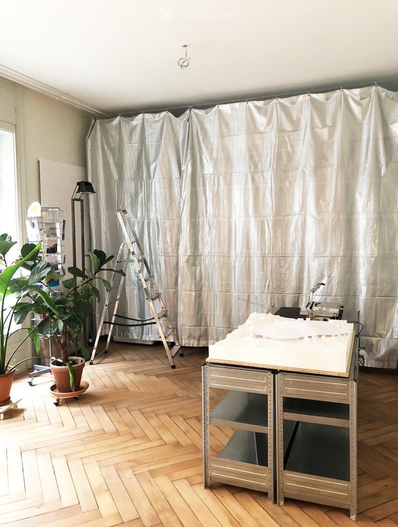 A room with wooden flooring, a large potted plant, a ladder, a table with a white object on it, and a lamp near the window.