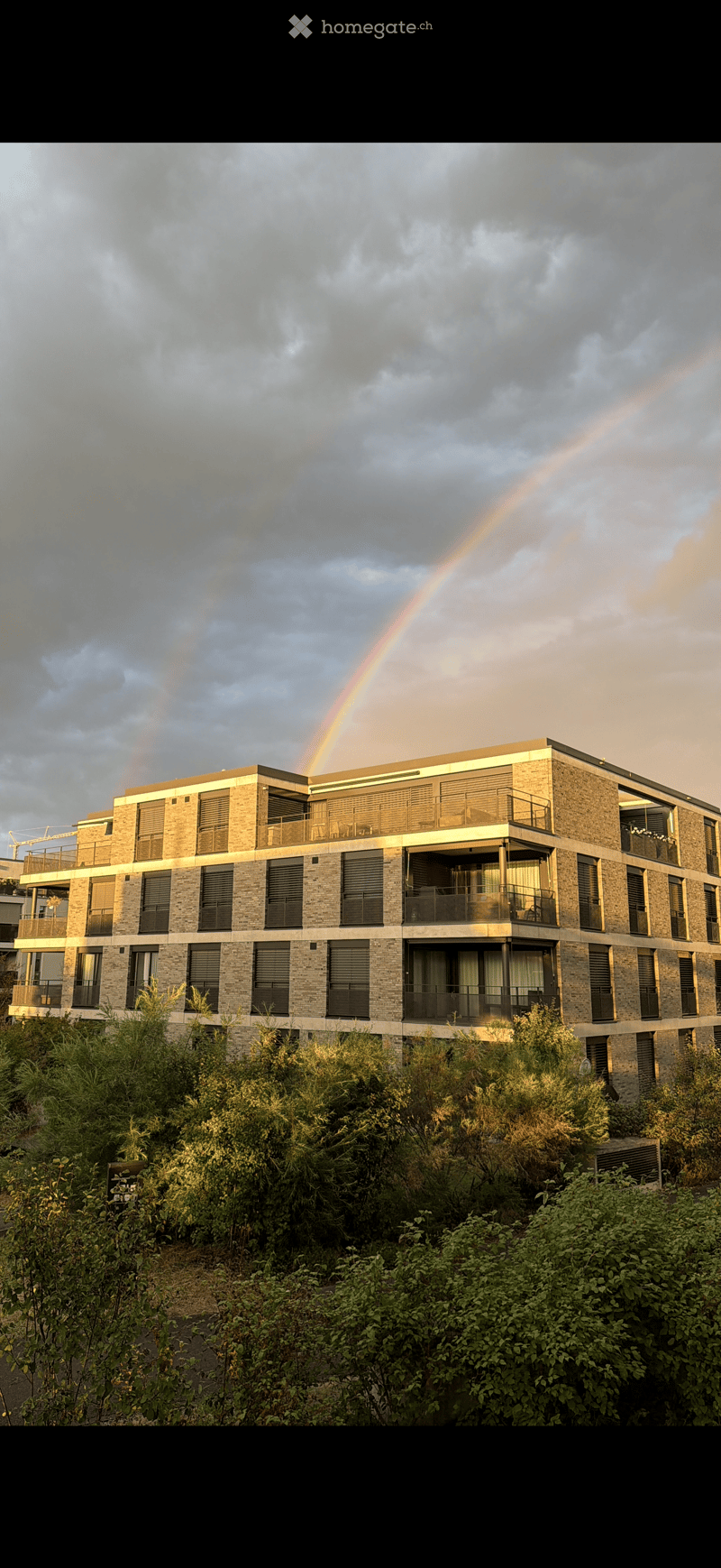 Modern multi-story residential building with brick facade, balconies, and large windows, surrounded by greenery with a visible rainbow in the sky.