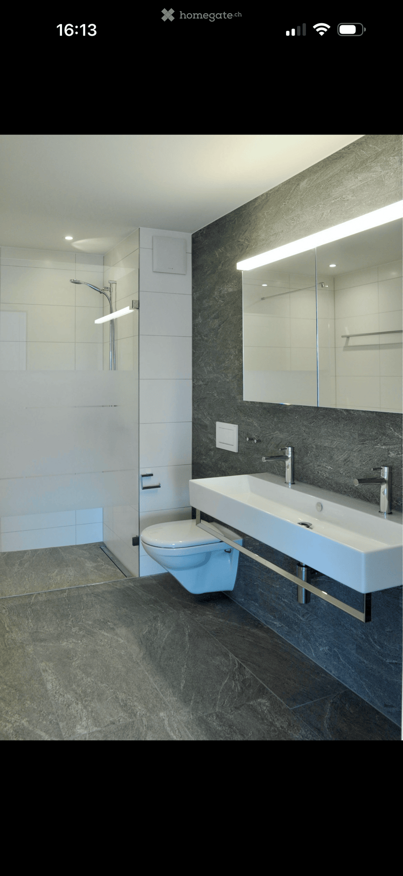 Modern bathroom with a large sink, toilet, and shower area. Features dark marble tiles on the walls and floor, a large mirror with built-in lighting, and sleek chrome fixtures.