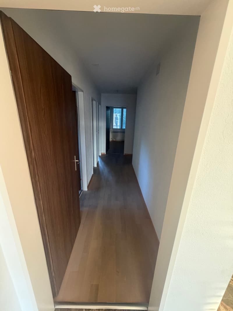 Empty hallway with wooden flooring, white walls, and a partially open wooden door on the left side.