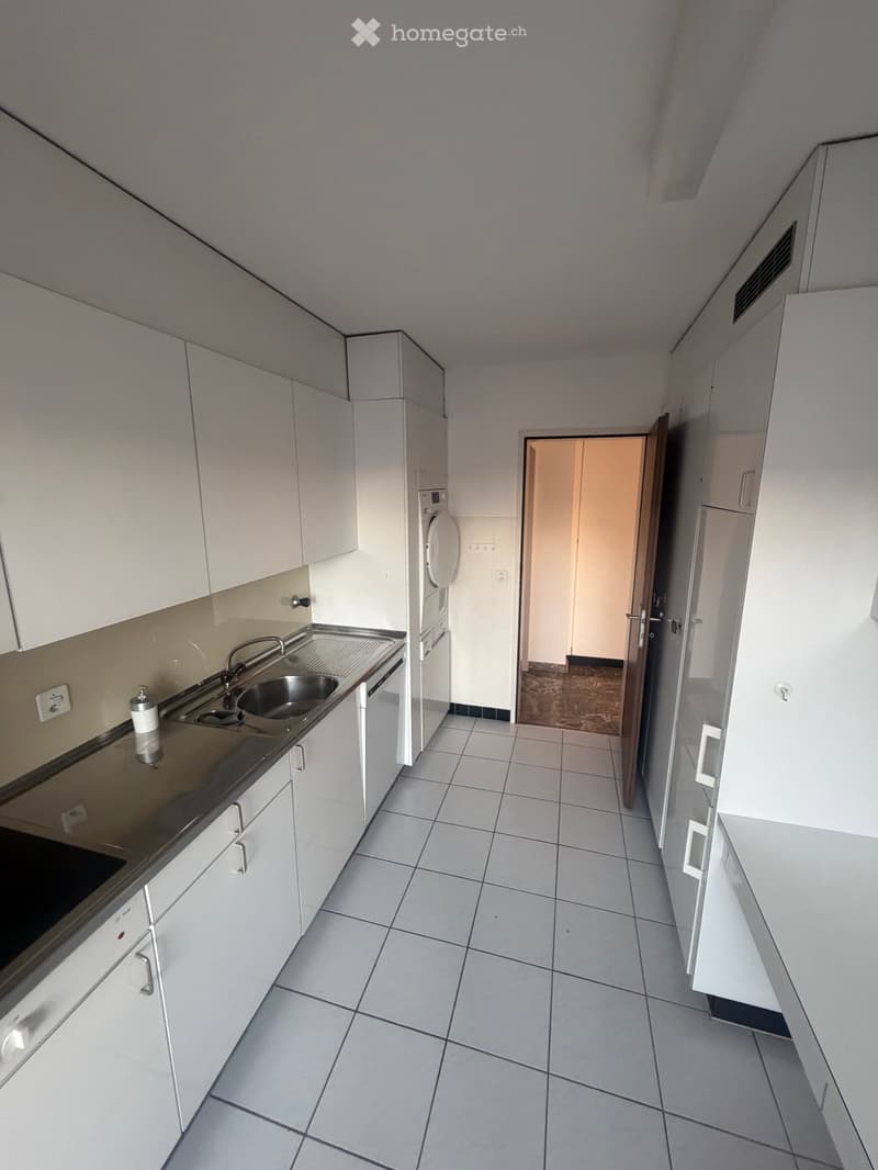 Empty kitchen with white cabinets, tiled floor, and stainless steel sink. A doorway leads to another room.