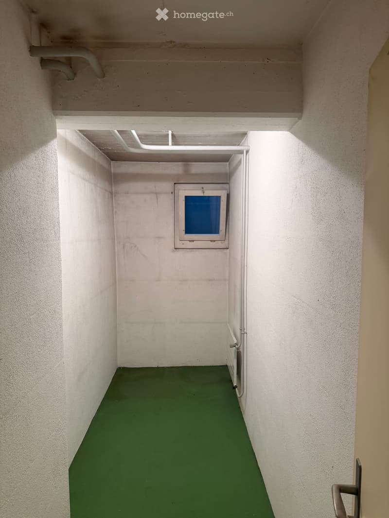 A narrow, green-floored corridor with white walls, a small window, and visible pipes along the ceiling and walls.