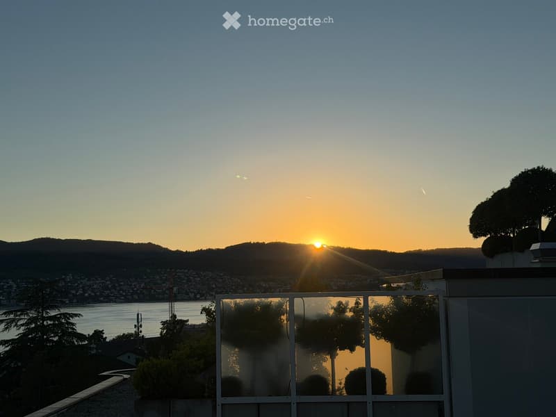 Balcony with a glass railing, overlooking a scenic sunset view of a city and lake.