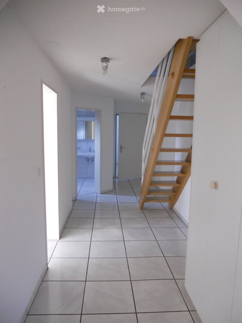 Empty hallway with tiled floor, white walls, wooden staircase leading upwards, and a view into a bathroom area.