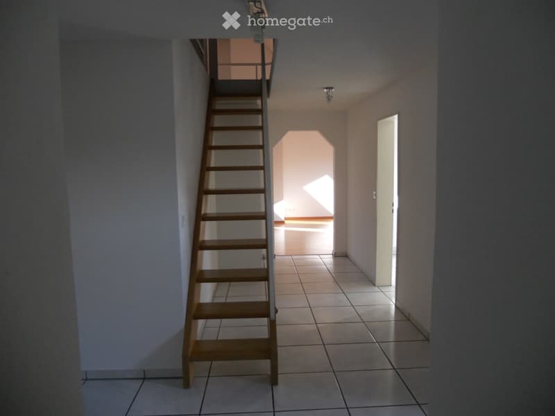 Empty hallway with tiled floor, wooden staircase leading upwards, and a partially visible adjacent room.