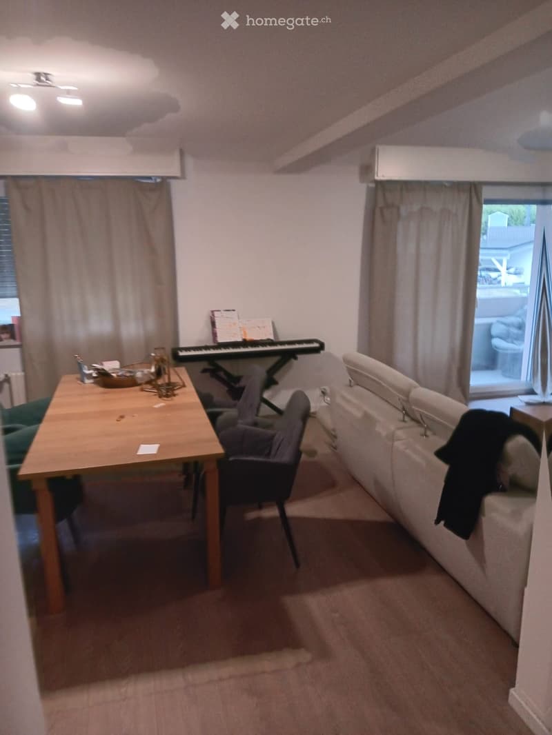 A cozy living room with a sofa, a wooden table, a chair, and a keyboard. The room has carpeted flooring, large windows with curtains, and a slanted ceiling typical of an attic space.