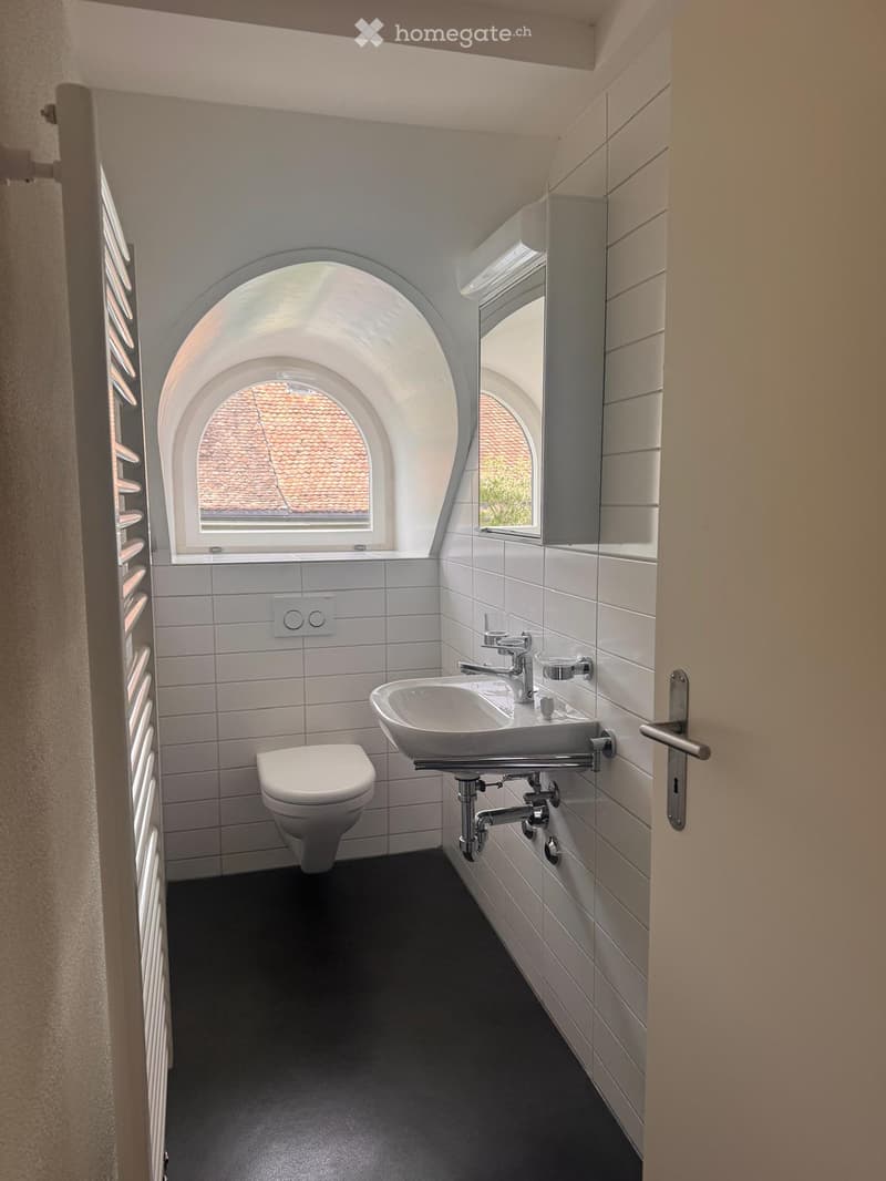 Modern bathroom with white tiles, black floor, toilet, sink, and mirror. Features arched windows and sleek fixtures.