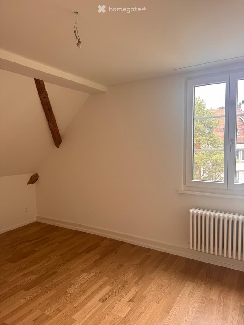 Empty attic room with wooden flooring, white walls, and a large window showing a view of trees and neighboring buildings.