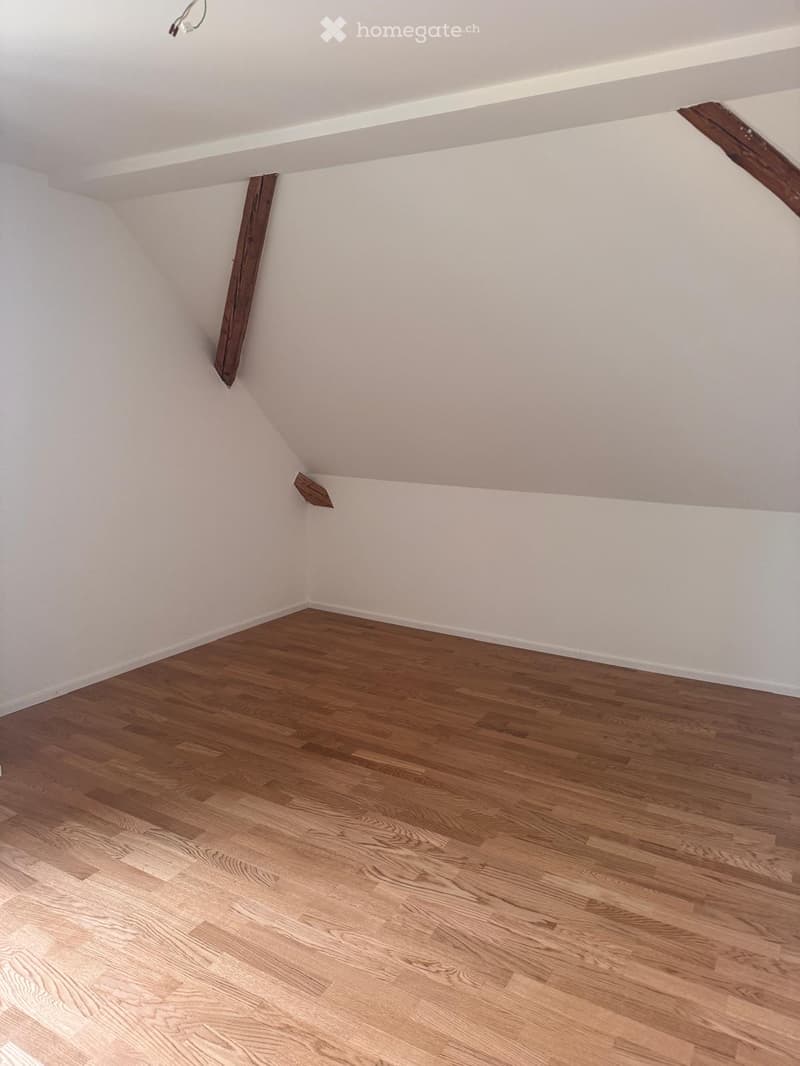 Empty attic room with wooden flooring and white walls, featuring exposed wooden beams in the ceiling.