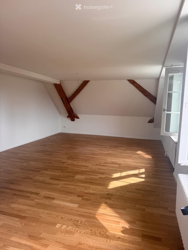 Empty room with wooden flooring, white walls, and exposed wooden beams on the ceiling. Sunlight streams in from a window on the right side.
