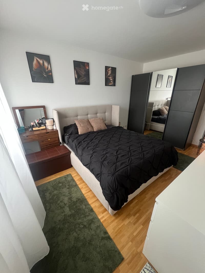 Modern bedroom with a large bed, nightstand, mirror, wardrobe, and a small desk area with a lamp and stool.