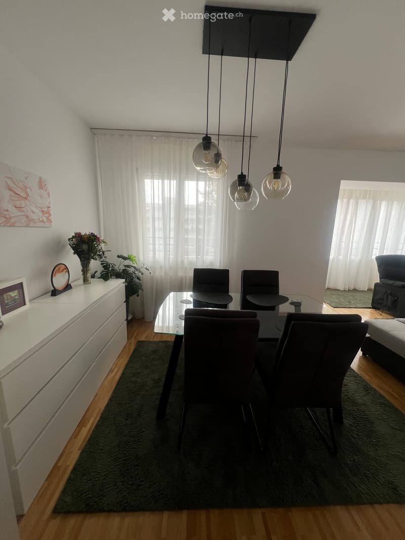 Modern living and dining area with wooden flooring, large windows with white curtains, a glass dining table with four chairs, a sofa, a white cabinet with a vase and plant, and pendant lights.