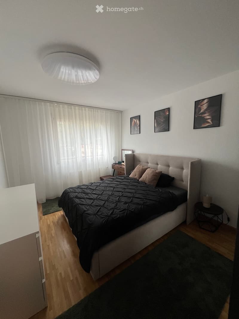 Modern bedroom with a large bed, nightstand, mirror, and artwork on the walls. The room features wooden flooring and large windows with white curtains.